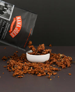 Nana Joes Granola Dark Chocolate blend pouring into a white bowl on a two-tone black background.